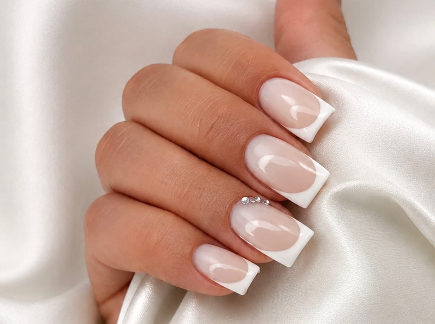 French manucure blanche sur ongles carrés, base nacrée et trois strass alignés, main posée sur de la soie ivoire